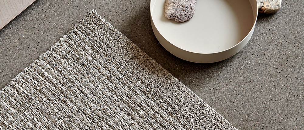 Close-up of the Pappelina Sam dark linen rug, highlighting its woven texture on a polished concrete floor. The image also features a decorative plate with a stone and a larger stone placed next to it.